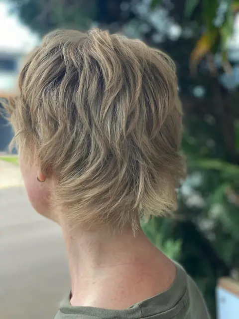 Back view of a choppy textured blonde pixie mullet, outdoors with greenery