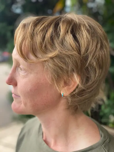 Left profile of a short golden strawberry blonde textured pixie cut, garden background